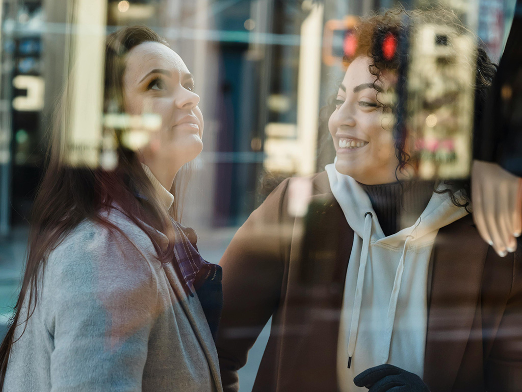 Two women stand in front of a shop window and laugh